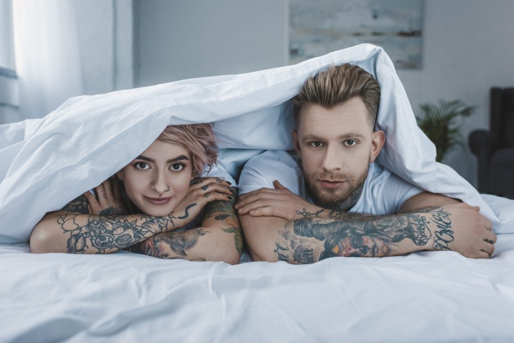 young tattooed couple lying in bed under white blanket